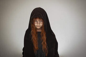 Halloween portrait of a small ominously smiling red-haired witch with loose hair dressed in a black...