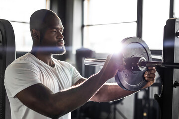 Muscular sporty african man adding weight on barbell at gym.
