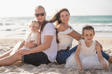 Fototapeta premium Summer vacation, happy family of four having fun. parents and two daughters sit on beach sea in summer day. white clothes and jeans. 