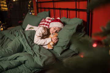 Fototapeta na wymiar a little girl in a santa hat sleeps on a green bedding in her bedroom for christmas, christmas magic, a tired child sleeps after celebrating new year or christmas