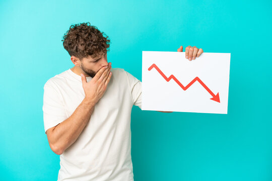 Young handsome caucasian man isolated on blue background holding a sign with a decreasing statistics arrow symbol with surprised expression