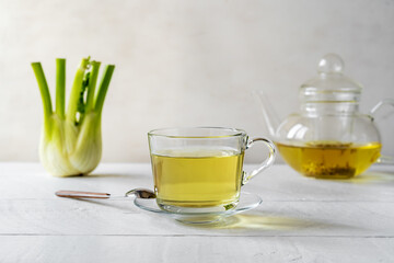 Fennel tea in a glass cup, fresh fennel bulb, seeds and tea pot on white wooden table with grey background