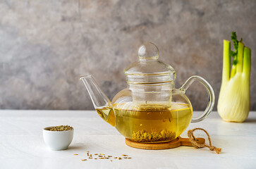 Fennel tea in a glass teapot, fresh fennel bulb, fennel seeds on white wooden table with grey background