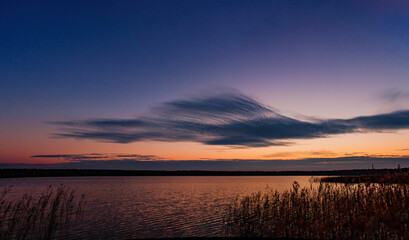 Calm light autumn sunset on the shore of the lake. With a plume cloud. In blue and red tones. The edge of the lake with a forest on the horizon.