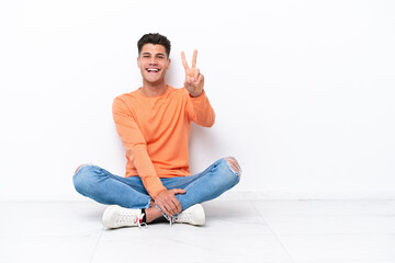 Young man sitting on the floor isolated on white background smiling and showing victory sign