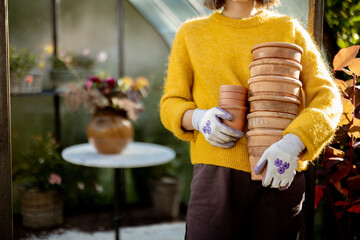 Woman stands with a pile of clay jugs in front of glass orangery at garden in sunny morning. Close-up on hands - obrazy, fototapety, plakaty