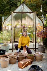 Woman plants flowers in clay jugs while sitting in front of beautiful glass orangery at garden. Florist gardening at beautiful backyard on sunny morning