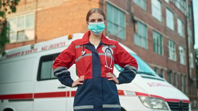 One young beautiful woman paramedic standing outdoor wearing medical mask and looking at camera near ambulance vehicle. Emergency worker. Professional doctor or nurse at work. First aid assistance
