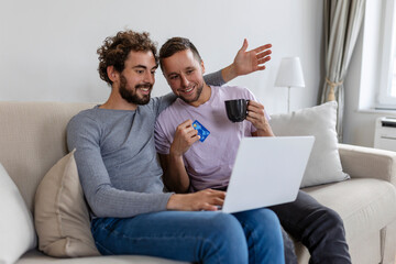 Cheerful young gay couple smiling cheerfully while shopping online at home. Two young male lovers using a credit card and a laptop to make a purchase online. Young gay couple sitting together indoors.