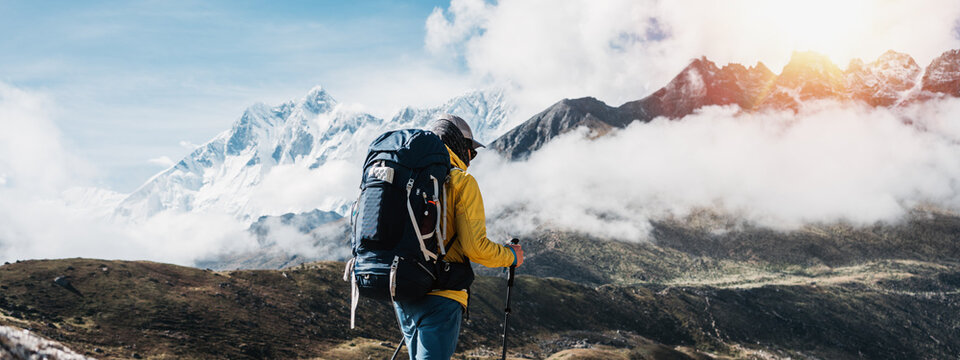 Outdoor tourist traveling along high altitude mountains wearing yellow jacket and professional backpack. Young solo hiker walk across sunny mountain track. Wide image