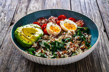 Boiled eggs with avocado, boiled red and white rice, cherry tomatoes, spinach and parmesan on wooden table
