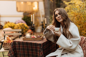 The girl drinks coffee tea in the garden against the backdrop of a campervan. Picnic in nature in autumn