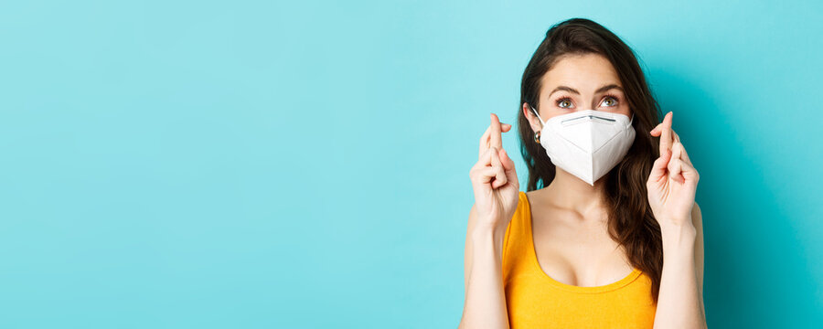 Health, Covid-19 And Lockdown Concept. Close Up Of Hopeful Young Woman In Respirator Mask, Cross Fingers And Making Wish, Praying And Looking Up, Blue Background