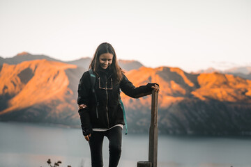 young caucasian brunette woman with loose hair black jacket black pants and glasses hanging from her neck smiling happy and content leaning on a wooden post at sunset between the mountains, roys peak