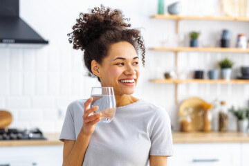 Healthy lifestyle concept. Beautiful young African American woman holding glass of fresh clean water standing in the home kitchen, looking at away and smiles friendly - Powered by Adobe