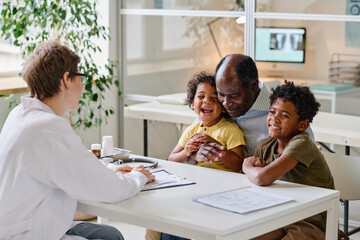 Happy little children sitting together with their dad at table and talking to pediatrist at office