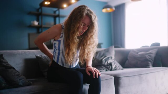 Caucasian woman sitting down on a couch at home experiences sudden back pain. Young adult female suffers from pain in her back descending on sofa. Girl rubbing back suffering from cramps in lumbar
