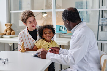 Young woman visiting pediatrician with her little son, they sitting at table and talking to doctor