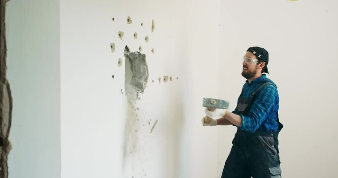 A smiling guy demolishes a wall in a house using a construction hammer on a long stick. Fun demolition of an apartment during renovation, getting rid of an unnecessary wall gaining space.