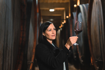 Woman in the wine cellar with barrels in background drinking and tasting wine. - obrazy, fototapety, plakaty
