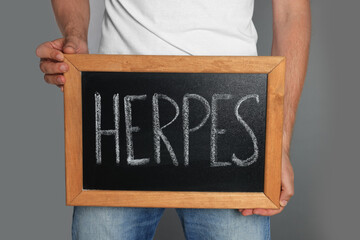 Man holding small chalkboard with word Herpes on grey background