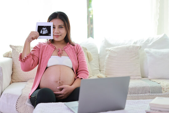 Happy smiling young asian pregnant woman resting and sitting on sofa in living room while showing ultrasound image with computer. Expectant mother preparing and waiting for baby birth during pregnancy