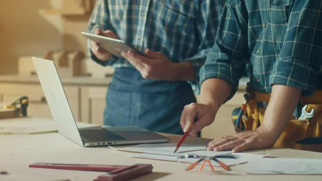 Small Business Owners of a Furniture Workshop Using Tablet, Notebook and Discussing the Design of a New Wooden Product. Hands of Carpenter and Young Male Apprentice Working in Studio.