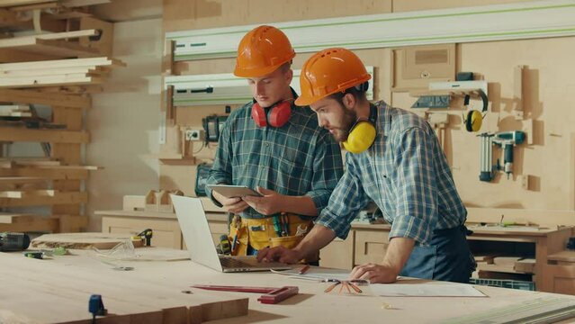 Two Carpenter Using Tablet, Notebook and Discussing the Design of New Wooden Product. Carpenter and Apprentice Working with Construction Noise Cancelling Headphones on their Neck and Hard Hat on Head