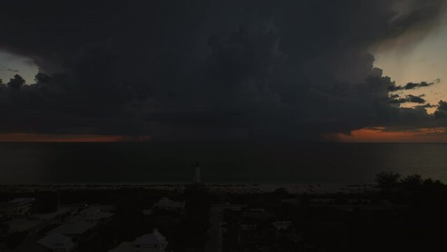 Seashore town with dark houses and tall lighthouse on ocean shore with blinking light at stormy night for vessels navigation. Thunderstorm with lightnings over sea water posing danger for ships