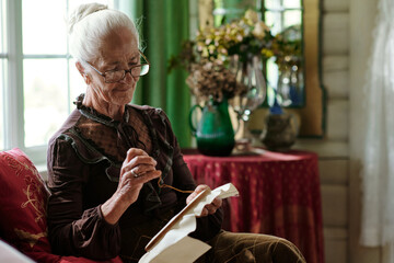 Pretty senior woman with canvas in hoop cross stitching at leisure while sitting on couch in living...