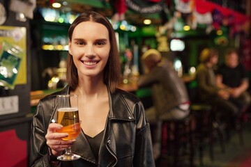 Photo realistic digital collage of pretty brunette girl in black leather jacket holding glass of beer while standing in front of camera in bar