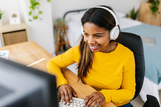 Cheerful teenage student girl with headphones learning online from home - Millennial hispanic latin woman studying on virtual platform website sitting at desk in bedroom - E-learning concept