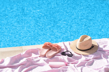Fototapeta na wymiar Pink blanket with slippers, hat, sunglasses and sunscreen near outdoor swimming pool on sunny day, space for text
