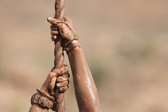 Mud race runner. Detail of muddy hands they climb the rope. Athletic man working out and climbing a rope, during obstacle course