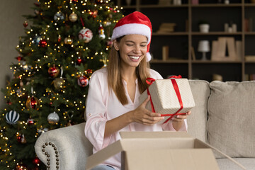 Joyful sincere young woman in festive hat taking wrapped gift from big carton box, feeling excited...