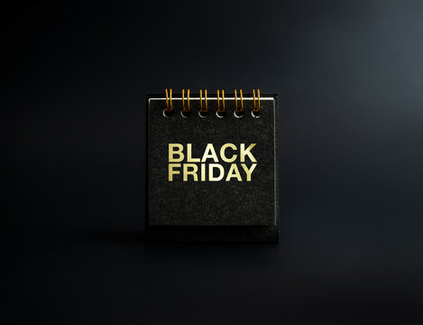 Black Friday sale concept. BLACK FRIDAY, gold text on small black spiral desk calendar cover standing on dark background, front view, minimal style. Invitation card, poster and banner.