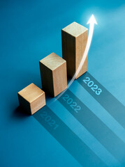 Shining rise up arrow on wooden cube blocks, bar graph chart steps on blue background with year...