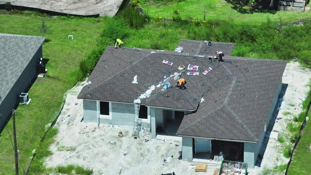 Top view of professional workers installing bitumen covering material on residential home wooden roof with asphalt shingles. House under construction in suburbs. Real estate development concept