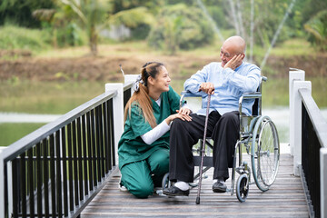 beautiful nurse and an elderly man on a wheelchair laughing happily on a bridge by the pool in the...