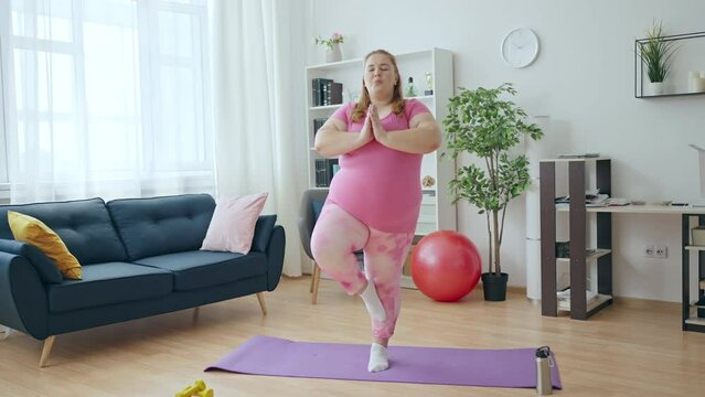Obese young woman training home, doing yoga exercise on one leg, meditating