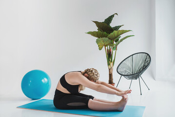 A plus size woman in a black sports top and leggings is doing morning exercises on a yoga mat. Chubby blonde is engaged in fitness. Curvy girl doing stretching in a bright room sitting on the floor