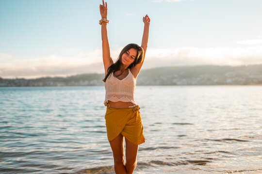 Happy young caucasian woman posing with arms up on the beach at sunset wearing a pareo skirt