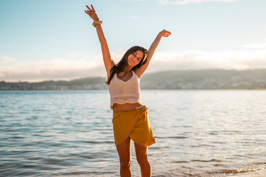 Happy young caucasian woman posing with arms up on the beach at sunset wearing a pareo skirt
