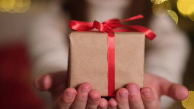 Kid hands giving small handmade Christmas gift box with red bow close up on background of glowing lights of Christmas tree. Person with present. Winter holidays, happy new year, celebration concept