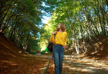 Young woman with backpack walks through the city forest park and enjoys nature on autumn sunny day