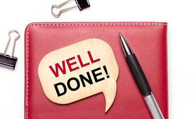 On a light background there are black paper clips, a pen, a burgundy notepad a wooden board with the text WELL DONE
