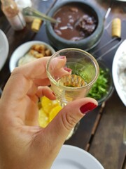 Brazilian pinga cup with feijoada meal background.  Hand holding cachaça from fermented sugarcane juice.