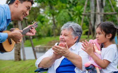 Fototapeta premium Happy disabled grandma having fun and clapping hands with her son and grandaughther playing ukulele.