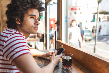 Thoughtful multiracial man drinking soda and using phone at cafe bar