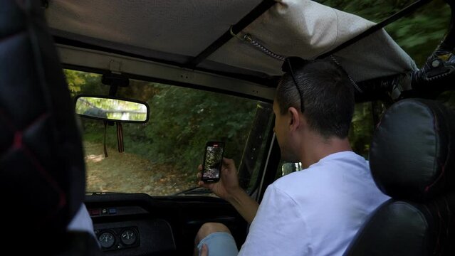 A Male Tourist Is Sitting In The Front Seat Of A Four-wheel Drive Jeep With A Phone, They Are Driving Along A Stone Road In The Mountains. Tourist Extreme Jeep Tour In The Mountains.